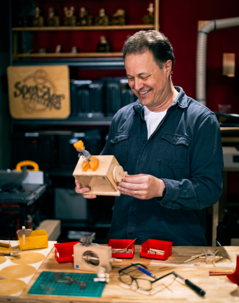 Gary Spetzler in his shop smiling and playing with his very first automata, a dancing (twirling) mouse with a large wedge of cheese above it's head.